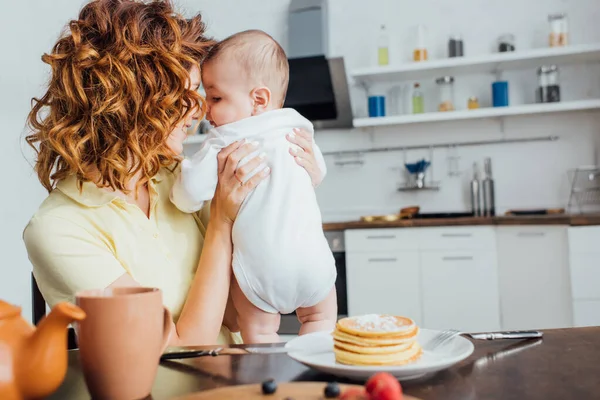 Enfoque selectivo de la madre joven rizado sosteniendo hijo cerca de la mesa servida con deliciosos panqueques - foto de stock