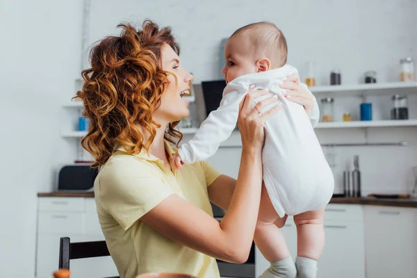 Young, curly mother holding infant boy in kitchen — Stock Photo