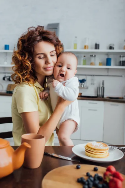 Messa a fuoco selettiva della mamma felice con gli occhi chiusi che tengono il bambino in pagliaccetti vicino al tavolo servito in cucina — Foto stock