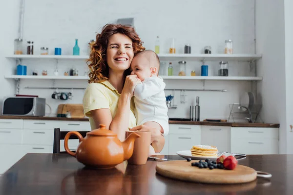 Enfoque selectivo de la madre joven sosteniendo niño en mamelucos cerca de la mesa servida con el desayuno - foto de stock