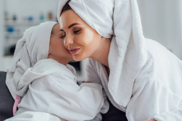 Daughter telling secret to young mother while sitting together in bathrobes and towels on heads — Stock Photo