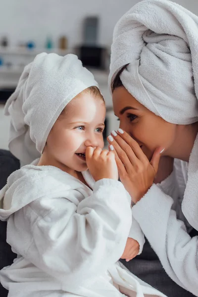 Jeune mère couvrant la bouche tout en disant secret à sa fille tout en étant assis ensemble dans des peignoirs et des serviettes sur la tête — Photo de stock