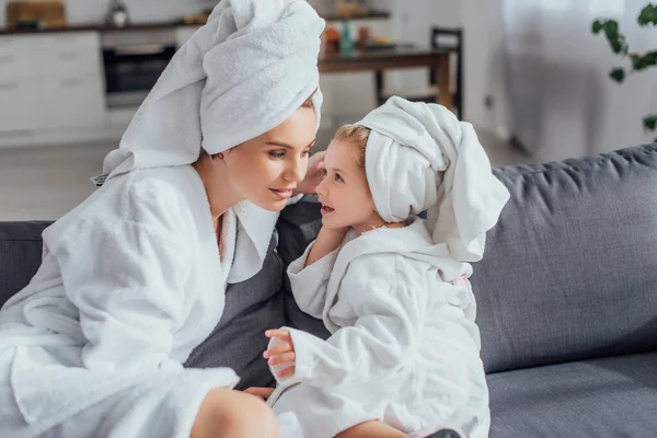 Selective focus of mother and daughter in white bathrobes and towels on heads talking while sitting on sofa — Stock Photo