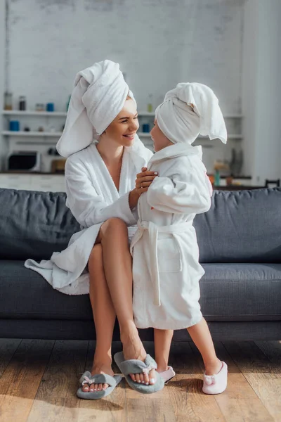 Young woman sitting on sofa and touching child in white bathrobe and towel on head — Stock Photo