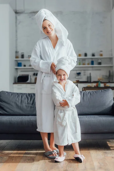 Молода мати і дочка в білих махрових халатах і рушниках на головах дивиться на камеру, стоячи біля дивана — стокове фото