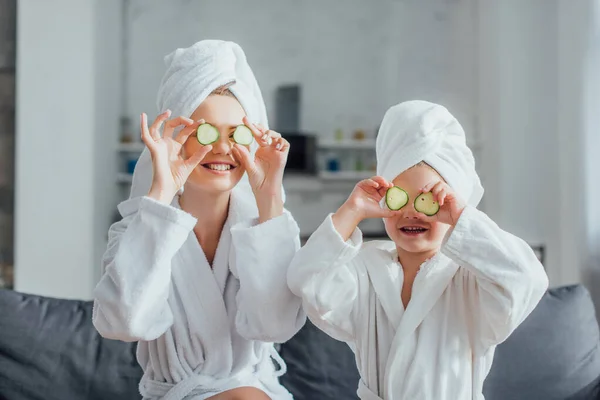 Young woman and daughter in white bathrobes and towels on heads applying fresh cucumber slices on eyes — Stock Photo