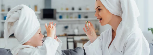 Side view of mother with child in white bathrobes and towels on heads holding fresh cucumber slices, panoramic concept — Stock Photo