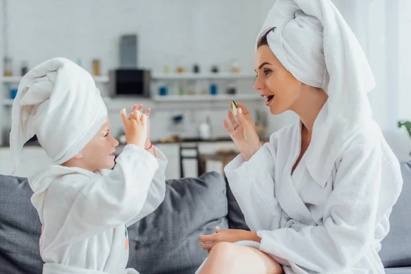 Боковой вид на молодую женщину с дочерью в белых халатах и полотенцах на головах, держащих свежие огуречные ломтики — стоковое фото
