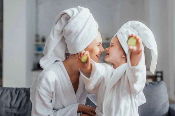 Mujer joven con hija en batas blancas y toallas en la cabeza sentada cara a cara mientras la niña sostiene rodajas de pepino fresco - foto de stock