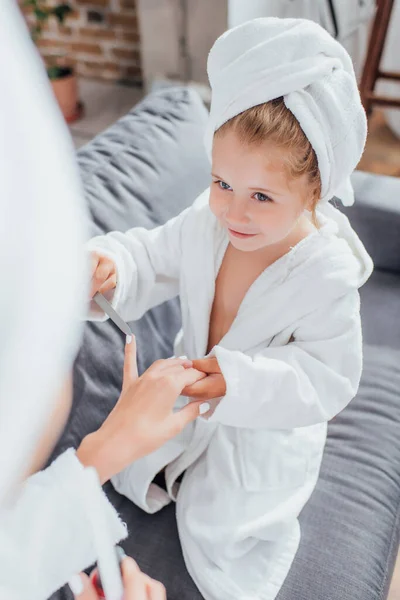 Selective focus of girl in white bathrobe and towel on head making manicure to mother — Stock Photo