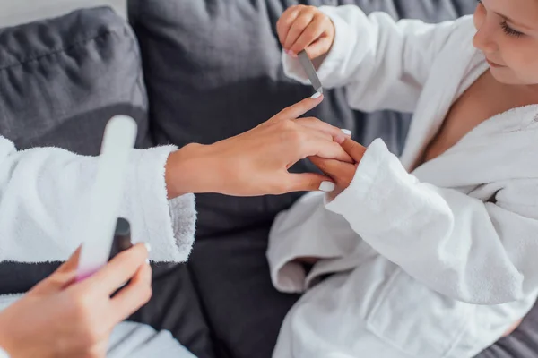 Vista recortada de la chica con la lima de uñas haciendo manicura a la madre mientras se sienta en el sofá en batas blancas juntos - foto de stock