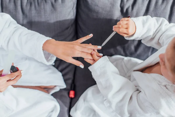 Vista dall'alto del bambino che fa manicure con lima per unghie alla madre mentre si siede sul divano in accappatoi bianchi insieme — Foto stock