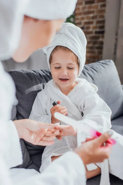 Селективное внимание женщины, держащей пилу для ногтей, делая маникюр дочери, сидящей на диване в белом халате — стоковое фото