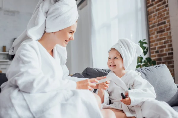 Selective focus of young woman making manicure to child while sitting together in white bathrobes and towels on heads — Stock Photo