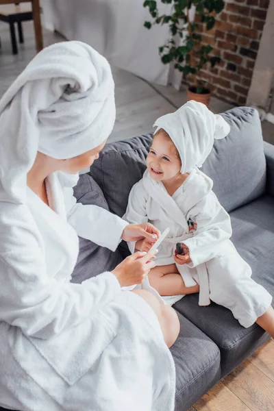 Высокий угол зрения женщины, делающей маникюр с пилкой для ногтей к дочери, сидя вместе в белых халатах — стоковое фото