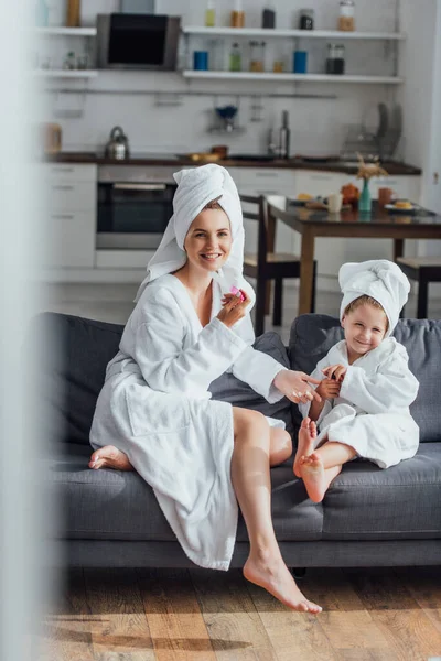 Foyer sélectif de la jeune femme tenant lime à ongles tout en étant assis sur le canapé avec sa fille en éponge peignoirs et serviettes sur la tête — Photo de stock