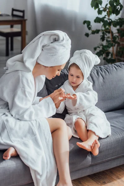 Молодая женщина делает маникюр дочери, сидя в белых халатах и полотенцах на головах вместе — стоковое фото