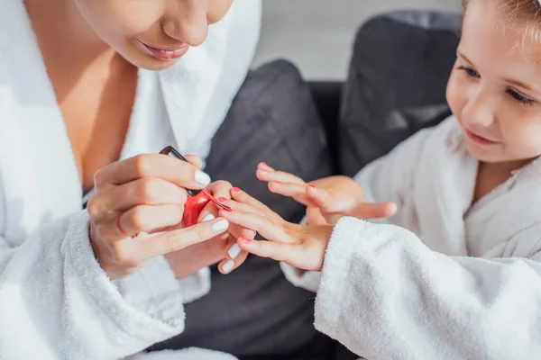 Vista cortada da mulher aplicando esmalte vermelho nas unhas da criança em roupão branco — Fotografia de Stock