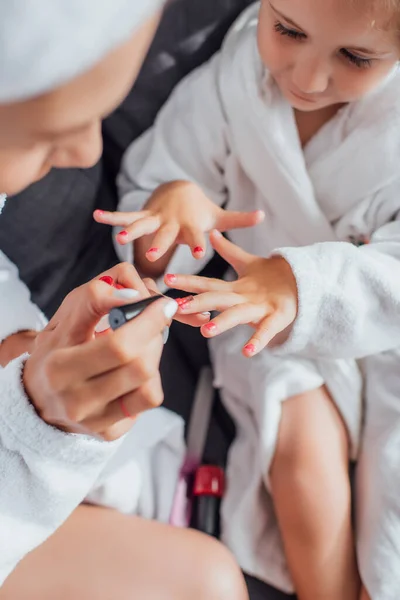 Vista de ángulo alto de la mujer aplicando esmalte rojo en las uñas de la hija en albornoz blanco - foto de stock