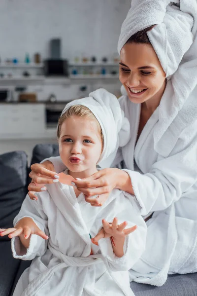 Junge Frau im weißen Bademantel mit Lippenstift auf Tochter sitzend mit ausgestreckten Händen — Stockfoto
