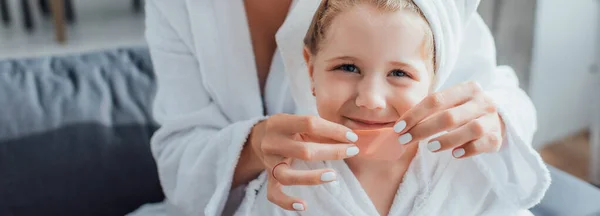 Cropped view of woman applying hydrogel patch on lips on child in white towel on head, horizontal image — Stock Photo