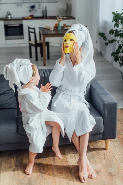 Visão de alto ângulo da mulher colocando máscara facial enquanto sentado com a filha em roupões de banho e toalhas na cabeça — Fotografia de Stock