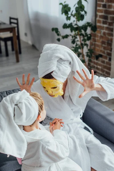 Vista de ángulo alto de la madre en la máscara facial que muestra gesto de miedo mientras está sentado con su hija en batas de baño y toallas en la cabeza - foto de stock