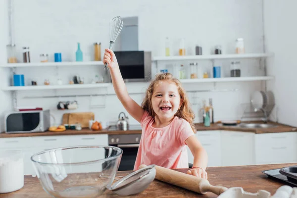 Kid holding whisk in raised hand near rolling pin, glass bowl and sieve on kitchen table — Stock Photo
