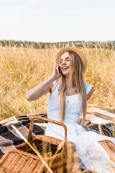 Selective focus of excited woman in white dress and straw hat talking on smartphone near wicker — Stock Photo