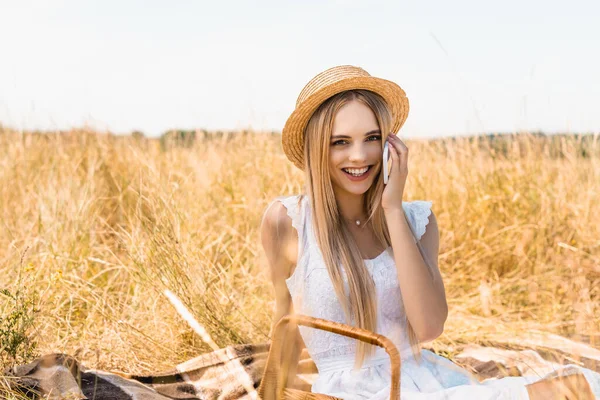 Selektiver Fokus einer stilvollen blonden Frau im Sommeroutfit, die auf dem Smartphone spricht, während sie auf dem Feld in die Kamera schaut — Stockfoto