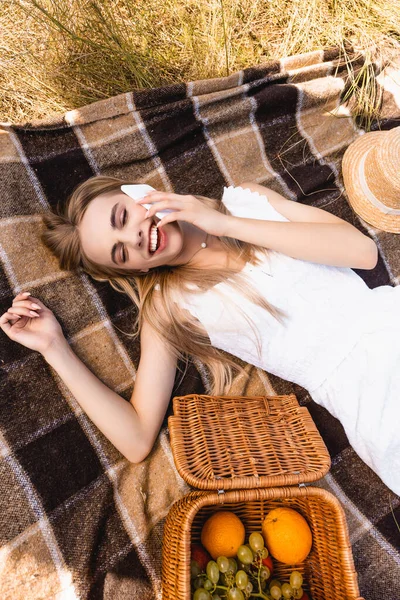 Top view of blonde woman in white dress lying on blanket near wicker basket with fruits and talking on smartphone — стоковое фото