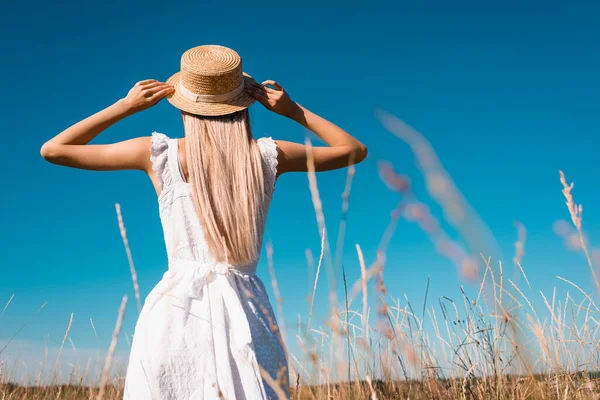 Back view of woman in white dress touching straw hat while standing in field against blue sky, selective focus — Stock Photo