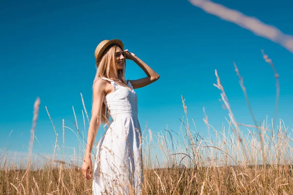 Foco seletivo da jovem mulher elegante em vestido branco tocando chapéu de palha e olhando para longe contra o céu azul — Fotografia de Stock