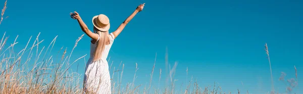 Vue arrière de la femme en robe blanche et chapeau de paille debout avec les mains tendues contre le ciel bleu, vue panoramique — Photo de stock