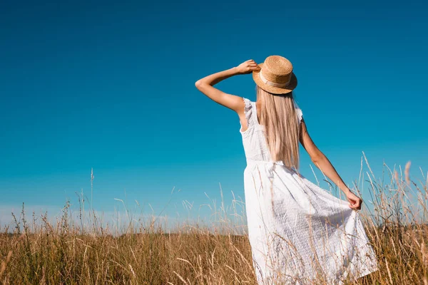 Vue arrière de la femme en robe blanche touchant robe blanche et chapeau de paille tout en se tenant debout dans prairie herbeuse — Photo de stock