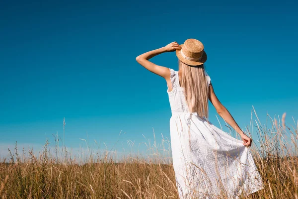 Rückansicht einer Frau in weißem Kleid, die weißes Kleid und Strohhut berührt, während sie auf einer Wiese steht — Stockfoto