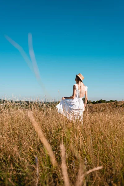 Вид ззаду стильна жінка в солом'яному капелюсі, що торкається білої сукні, стоячи на лузі на фоні блакитного неба, вибірковий фокус — стокове фото