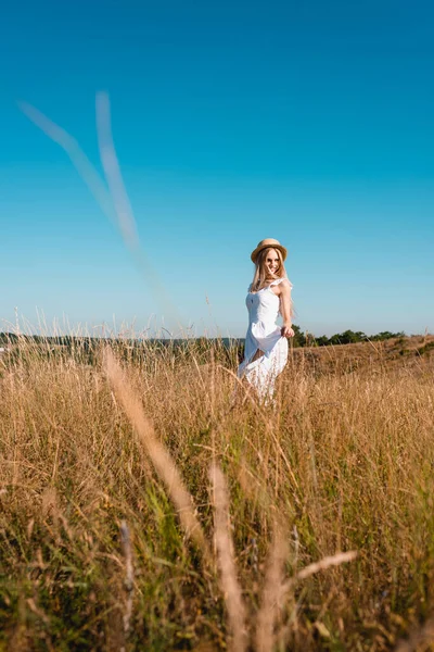 Вибірковий фокус стильної жінки в білій сукні і солом'яному капелюсі, що стоїть в полі на тлі блакитного неба — стокове фото