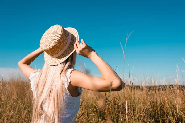 Vue arrière de la femme blonde en tenue d'été touchant chapeau de paille dans prairie herbeuse contre le ciel bleu, mise au point sélective — Photo de stock
