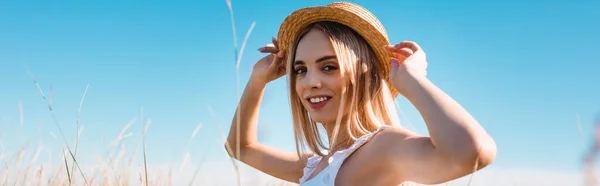 Selective focus of young blonde woman touching straw hat and looking at camera against blue sky, panoramic concept — Stock Photo