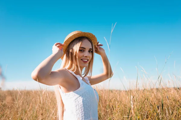 Селективный фокус чувственной блондинки, трогающей соломенную шляпу и смотрящей на камеру в травянистом поле — стоковое фото