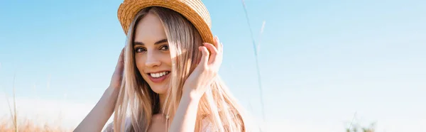 Horizontales Bild einer jungen blonden Frau, die Strohhut berührt, während sie vor blauem Himmel in die Kamera blickt — Stockfoto