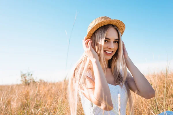 Селективный фокус блондинки, смотрящей в камеру и трогающей соломенную шляпу, сидя на солнце на фоне голубого неба — стоковое фото