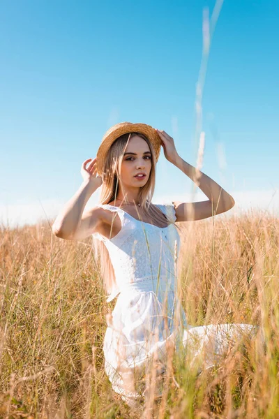 Selective focus of young blonde woman in white dress sitting in field and touching straw hat while looking at camera — Stock Photo