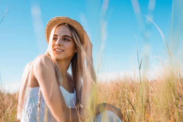 Вибірковий фокус стильної блондинки, дивлячись далеко, сидячи в полі і торкаючись солом'яного капелюха — стокове фото