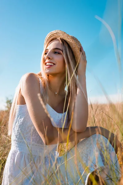 Selective focus of sensual blonde woman looking away while sitting in meadow and touching straw hat — Stock Photo