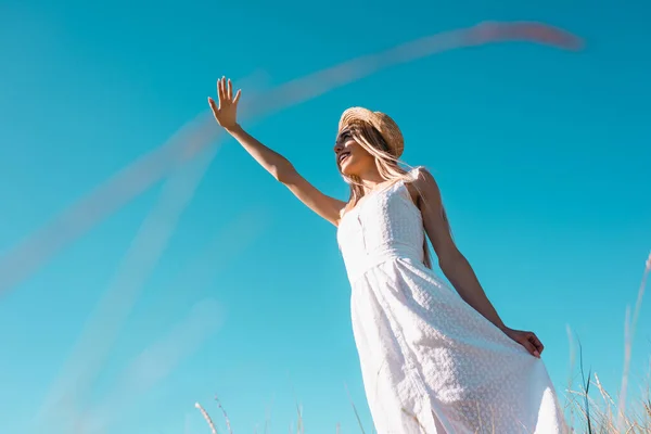 Baixo ângulo vista de mulher elegante em vestido branco acenando mão enquanto olha para longe contra o céu azul, foco seletivo — Fotografia de Stock