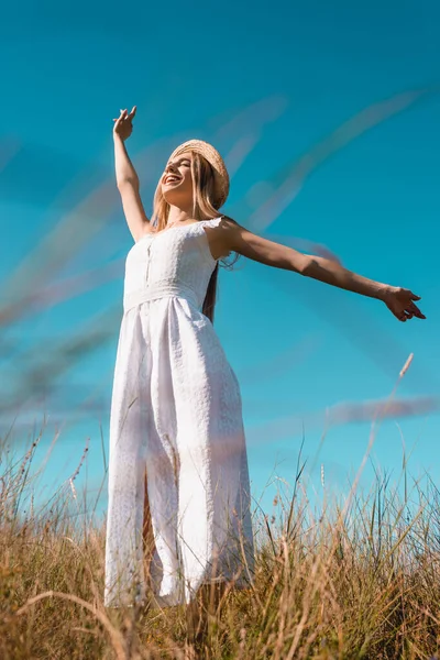 Tiefansicht einer stilvollen Frau im weißen Kleid, die mit ausgestreckten Händen und geschlossenen Augen vor blauem Himmel steht — Stockfoto