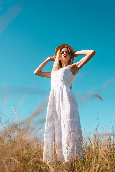 Selective focus of young woman in white dress touching straw hat while looking away against blue sky, low angle view — Stock Photo