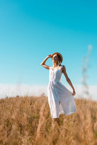 Messa a fuoco selettiva di donna elegante toccare abito bianco e cappello di paglia mentre in piedi in campo con gli occhi chiusi — Foto stock